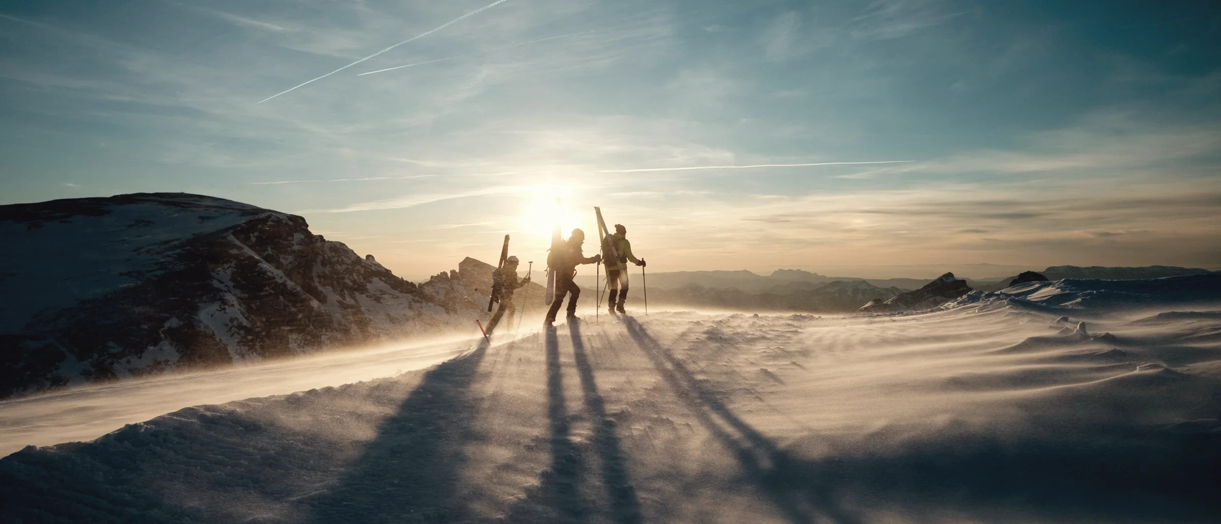 Three ski tourers silhouetted on a ridge against a low sun, spindrift blowing across the snow
